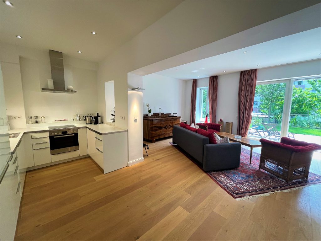 Offene Wohnküche im Apartmenthaus Christina in Nürnberg mit moderner Einbauküche, großzügigem Wohnbereich, Holzboden, stilvollem Mobiliar und Zugang zur grünen Terrasse – perfekte Kombination aus Komfort und Design nahe dem Design Hotel Vosteen. / Open-plan kitchen and living area at Apartmenthaus Christina in Nuremberg featuring a modern fitted kitchen, spacious lounge, wooden flooring, stylish furnishings and access to the green terrace – perfect blend of comfort and design near Design Hotel Vosteen.