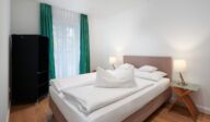 Schlafzimmer im Apartmenthaus Christina in Nürnberg mit gemütlichem Boxspringbett, pastellfarbenem Kopfteil, minimalistischer Einrichtung und grün-blauen Vorhängen – ideal für erholsame Nächte in zentraler Lage nahe dem Design Hotel Vosteen. / Bedroom at Apartmenthaus Christina in Nuremberg featuring a cozy box-spring bed, pastel headboard, minimalist furnishings and green-blue curtains – ideal for restful nights in a central location near Design Hotel Vosteen.