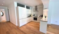 Moderne Einbauküche im Apartmenthaus Christina in Nürnberg mit weißen Hochglanzfronten, Holzfußboden, integrierten Geräten und stilvollem Lichtkonzept – ideal für Selbstverpflegung in zentraler Lage nahe dem Design Hotel Vosteen. / Modern built-in kitchen at Apartmenthaus Christina in Nuremberg featuring white high-gloss cabinets, wooden flooring, integrated appliances and stylish lighting – perfect for self-catering in a central location near Design Hotel Vosteen.