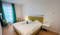 Schlafzimmer im Apartmenthaus Christina in Nürnberg mit komfortablem Doppelbett und senffarbenem Kopfteil, Holzboden, Retro-Stühlen und grünen Vorhängen – stilvoller Mix aus Vintage-Elementen und modernem Komfort nahe dem Design Hotel Vosteen. / Bedroom at Apartmenthaus Christina in Nuremberg featuring a comfortable double bed with mustard-colored headboard, wooden flooring, retro chairs and green curtains – stylish blend of vintage elements and modern comfort near Design Hotel Vosteen.