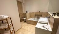 Hochwertig ausgestattetes Badezimmer im Apartmenthaus Christina in Nürnberg mit Badewanne, modernem Waschbecken, Natursteinfliesen, Regal mit Handtüchern und stilvollen Details – ideal für entspannte Auszeiten nahe dem Design Hotel Vosteen. / Well-equipped bathroom at Apartmenthaus Christina in Nuremberg featuring a bathtub, modern sink, natural stone tiles, towel rack and stylish details – ideal for relaxing moments near Design Hotel Vosteen.