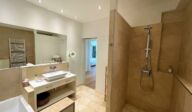 Modernes Badezimmer im Apartmenthaus Christina in Nürnberg mit begehbarer Dusche, großem Spiegel, Waschbecken im Waschtisch und hellen Natursteinfliesen – hochwertig ausgestatteter Wohlfühlbereich nahe dem Design Hotel Vosteen. / Modern bathroom at Apartmenthaus Christina in Nuremberg featuring a walk-in shower, large mirror, countertop washbasin and light natural stone tiles – high-quality wellness space near Design Hotel Vosteen.