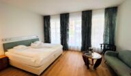 Helles Schlafzimmer im Apartmenthaus Christina in Nürnberg mit großem Doppelbett, modernen Vorhängen, gemütlicher Sitzecke und Holzboden – stilvoll eingerichteter Raum für einen komfortablen Aufenthalt nahe dem Design Hotel Vosteen. / Bright bedroom at Apartmenthaus Christina in Nuremberg featuring a large double bed, modern curtains, cozy sitting area and wooden flooring – stylishly furnished space for a comfortable stay near Design Hotel Vosteen.