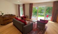Helles Wohnzimmer im Apartmenthaus Christina in Nürnberg mit direktem Zugang zur Gartenterrasse – stilvolle Einrichtung mit Holzboden, Retro-Kommode, Sofa mit roten Kissen und großer Fensterfront mit Blick ins Grüne. / Bright living room at Apartmenthaus Christina in Nuremberg with direct access to the garden terrace – stylish interior with wooden flooring, vintage sideboard, sofa with red cushions, and large windows overlooking the greenery.