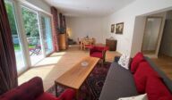 Großzügiger Wohn- und Essbereich im Apartmenthaus Christina in Nürnberg mit Zugang zur Gartenterrasse – gemütliches Sofa mit roten Kissen, Esstisch, antike Möbel und viel Tageslicht durch bodentiefe Fenster. / Spacious living and dining area at Apartmenthaus Christina in Nuremberg with access to the garden terrace – cozy sofa with red cushions, dining table, antique furniture and plenty of daylight through floor-to-ceiling windows.